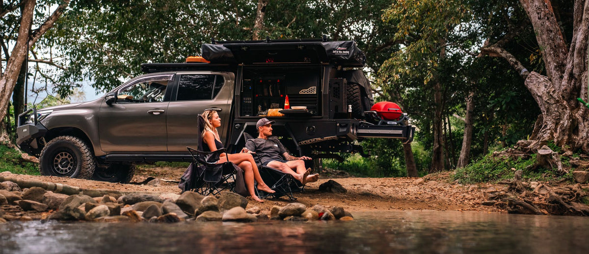 Toyota Hilux N80 with its XGO canopy open beside a creek, two people in camp chairs